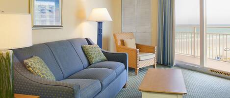 A hotel room with a blue sofa, a wooden coffee table, a wicker chair, a lamp, and a view of the beach through large windows.