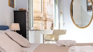 Une chambre d’hôtel comprenant un lit, un bureau, une chaise, une lampe, une armoire, un miroir et une fenêtre donnant sur un immeuble.