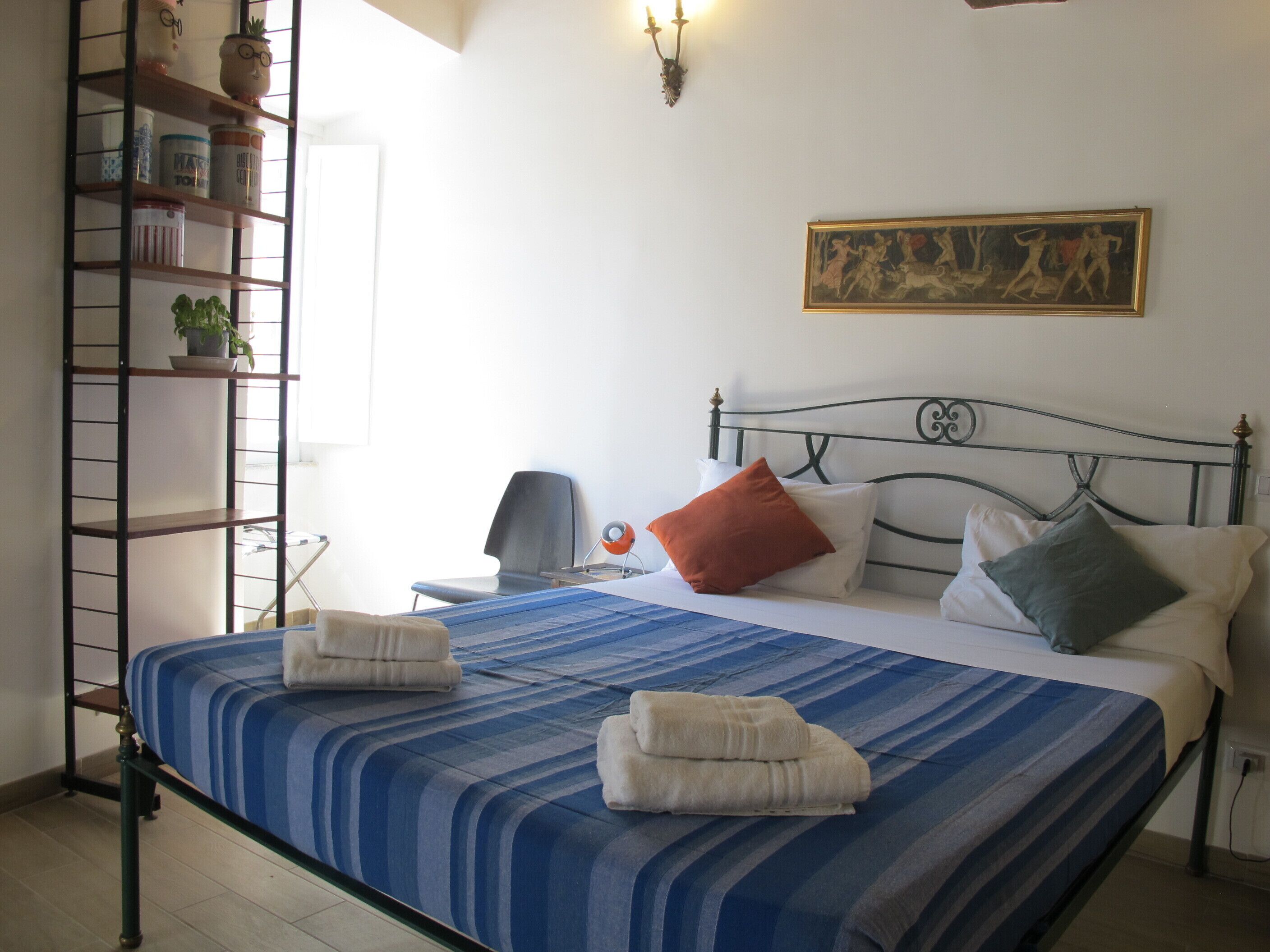 A bedroom with a bed, pillows, a nightstand, a lamp, a shelf with items, and a painting on the wall.