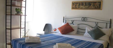 A bedroom with a bed, pillows, a nightstand, a lamp, a shelf with items, and a painting on the wall.