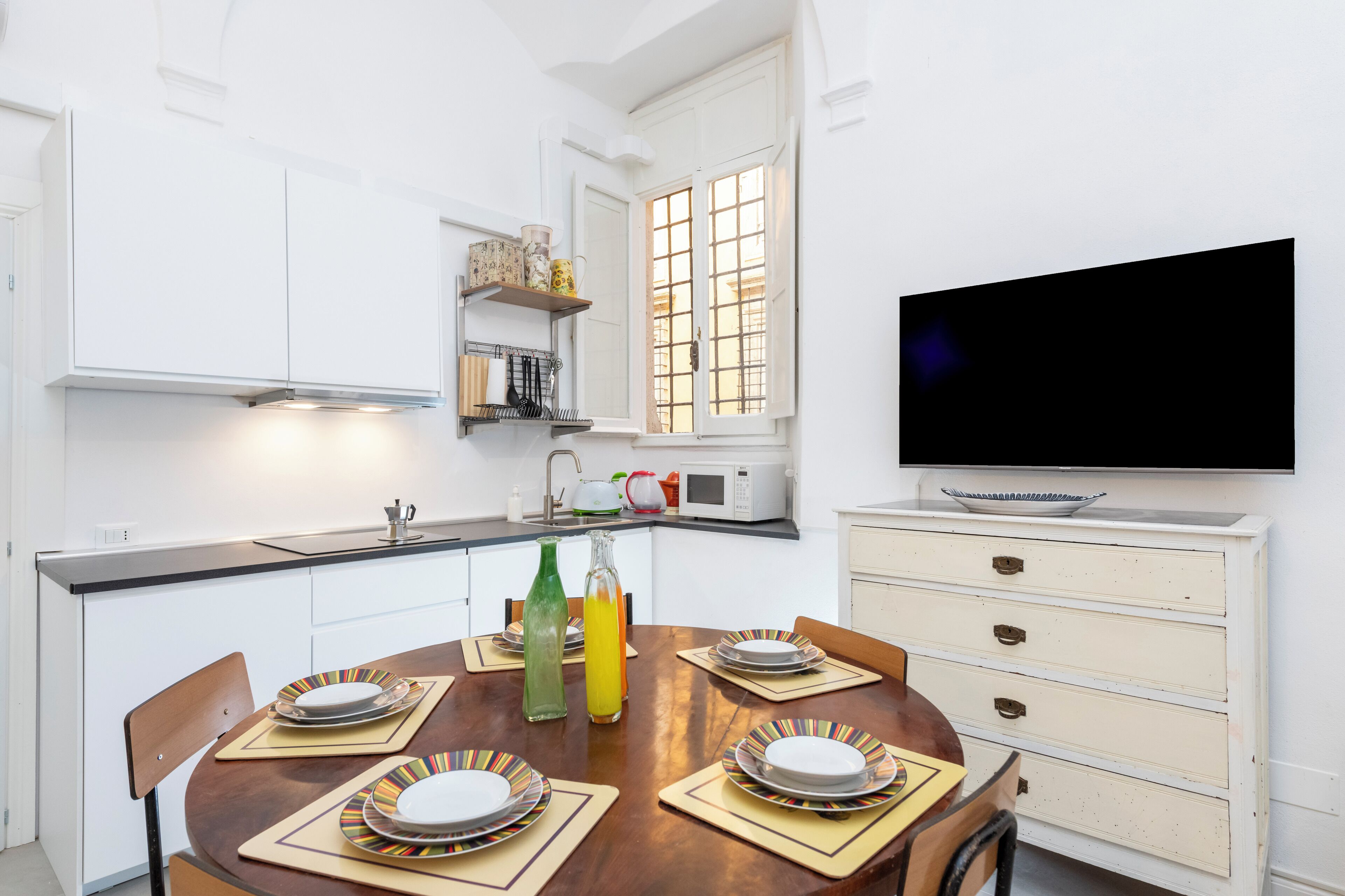 A dining area with a round table set for four, a television on the wall, and a kitchen in the background.