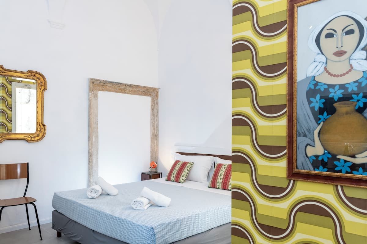 A hotel room with a bed, a chair, a nightstand, and a wall with a large mirror and a framed painting.
