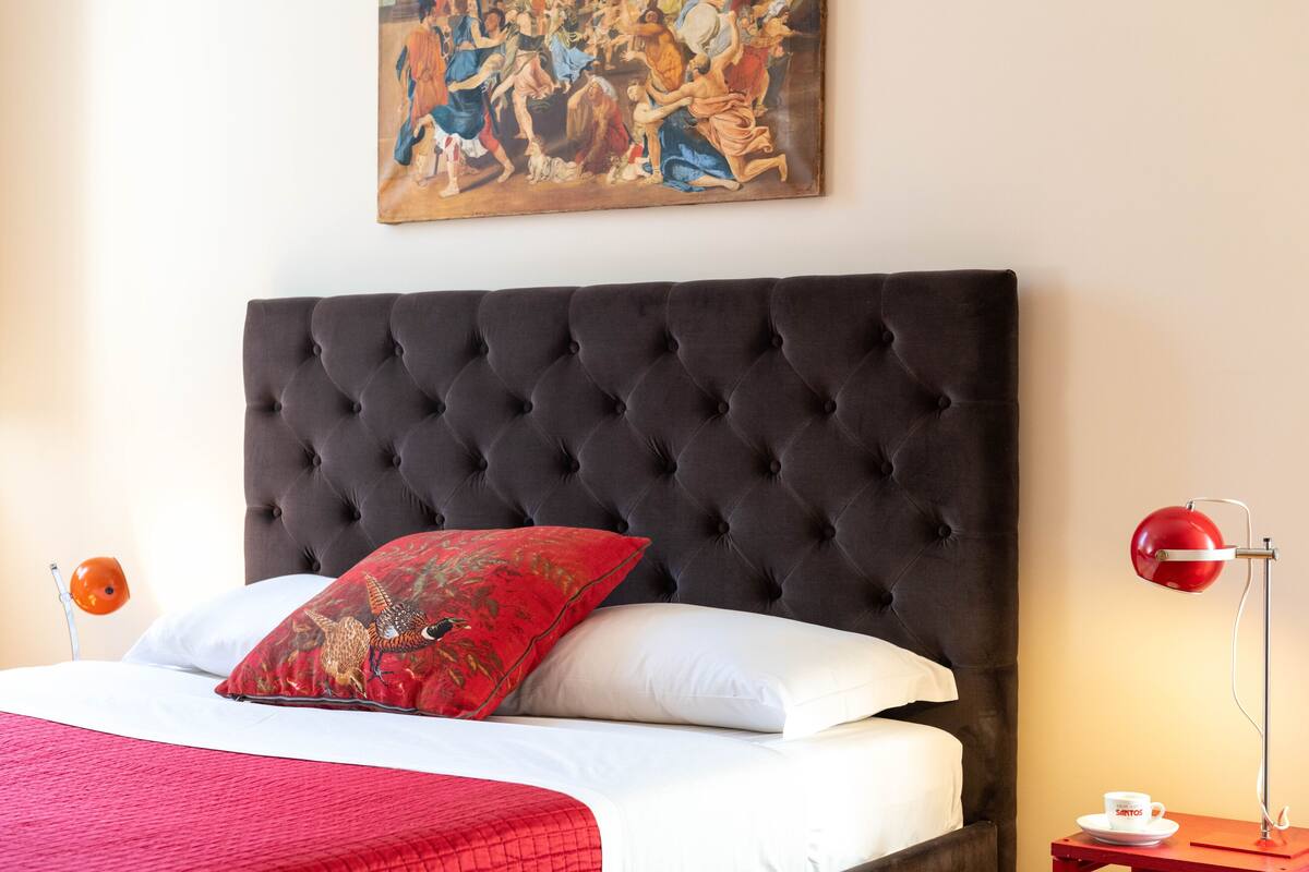 A neatly made bed with a tufted headboard, a red and white pillow, and a painting above it.