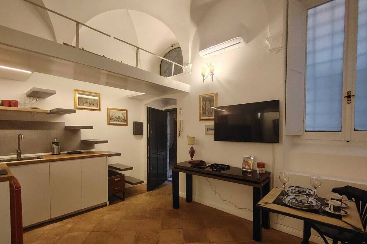A compact kitchen with a dining area, a TV mounted on the wall, and a window with blinds.