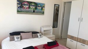 A neatly made bed with a pink bedspread and two red towels, a white chair, a wardrobe, and a painting of colorful houses on the wall.