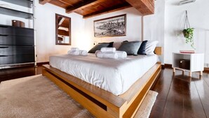 Un dormitorio moderno con una cama grande, vigas de madera, una planta colgante y un cuadro con una vista de la ciudad.