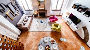 Amplia sala de estar con un sofá, una mesa de centro y un televisor. Cuenta con una chimenea, una mesa de comedor con platos y tazas, y una estantería.