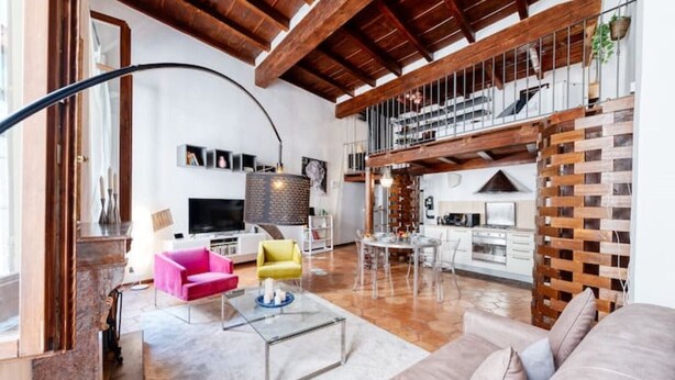 Amplia sala de estar con cocina moderna, vigas de madera y comedor con sillas traslúcidas.