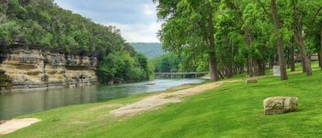 A riverside park with a grassy area, trees, and a bridge in the distance.
