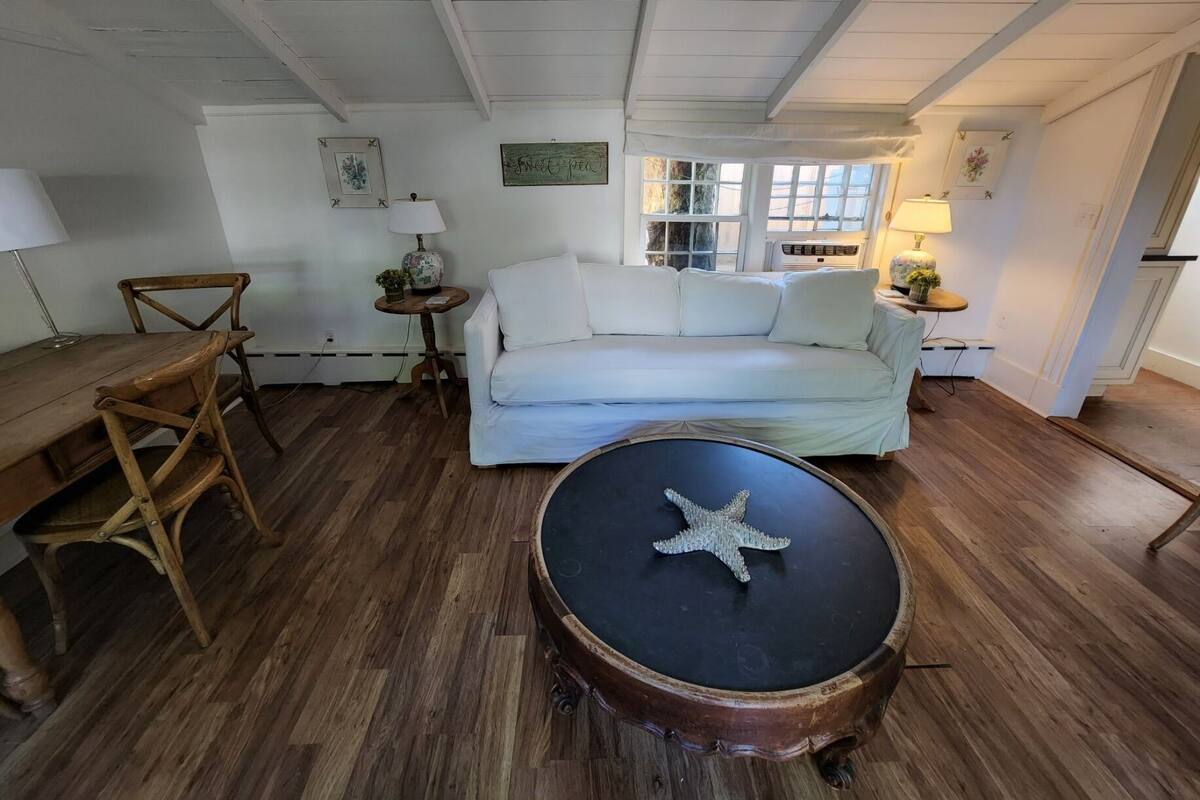 A cozy room with a wooden floor, a white sofa, a wooden chair, a round table with a starfish decoration, and a small side table with a lamp.