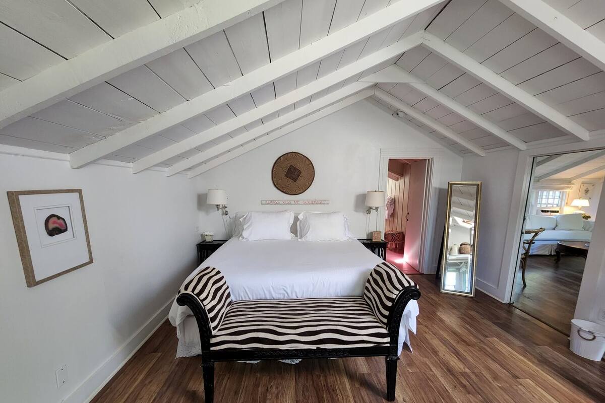 A bedroom with a high ceiling, a bed with a zebra-patterned throw, a round wall clock, and a mirror.