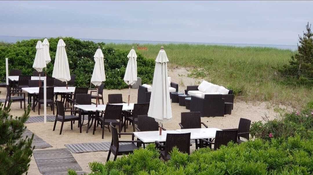 An outdoor dining area with tables and chairs, some covered by white umbrellas, set against a backdrop of greenery and a view of the sea.