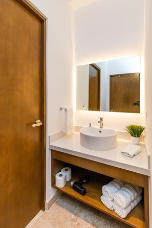 A bathroom with a wooden door, a white sink, a mirror, a small potted plant, and towels on the shelf. - Ipana By Riviera Rentals (Playa del Carmen)