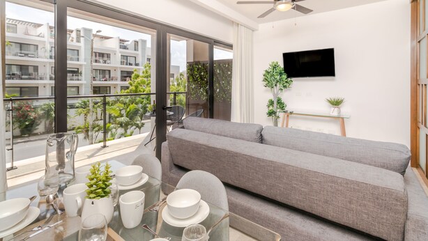A modern living room with a grey sofa, a glass dining table set with plates, cups, and cutlery, and a TV mounted on the wall. - Ipana By Riviera Rentals (Playa del Carmen)