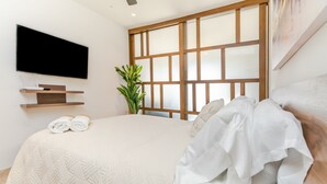 A modern bedroom with a large bed, a mounted TV, a plant, and a wooden headboard. - Ipana By Riviera Rentals (Playa del Carmen)