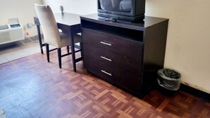 Ein Zimmer mit Holzboden, einem dunklen Holztisch mit Fernseher darauf, einem Stuhl, einer kleinen Mülltonne und einem Wandheizgerät.
