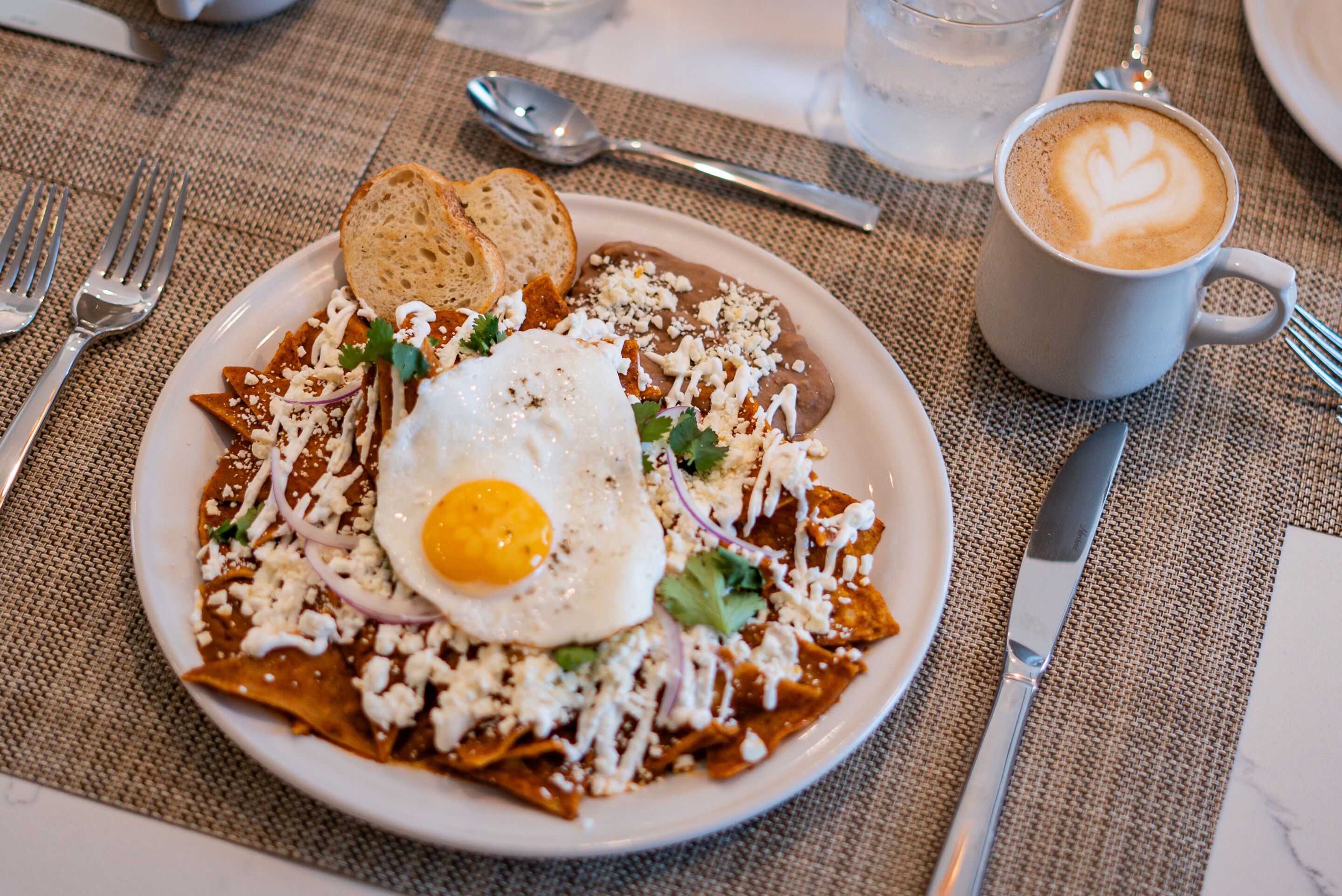 Een bord chilaquiles met een gebakken ei, kaas en zure room, vergezeld van een kop koffie met latte-art.