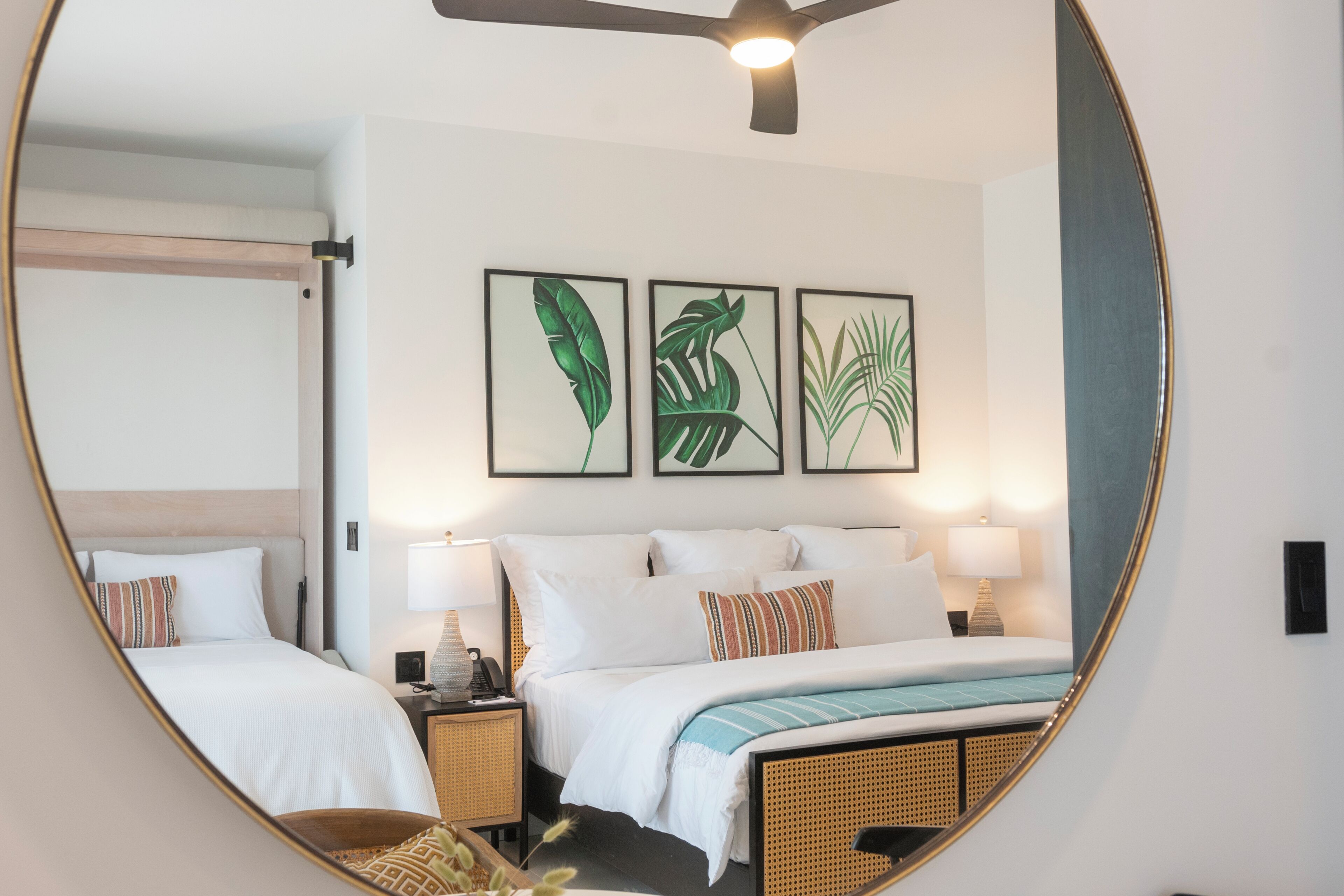 Habitación de hotel con cama, dos mesitas de noche, un espejo y tres cuadros con hojas enmarcadas en la pared.