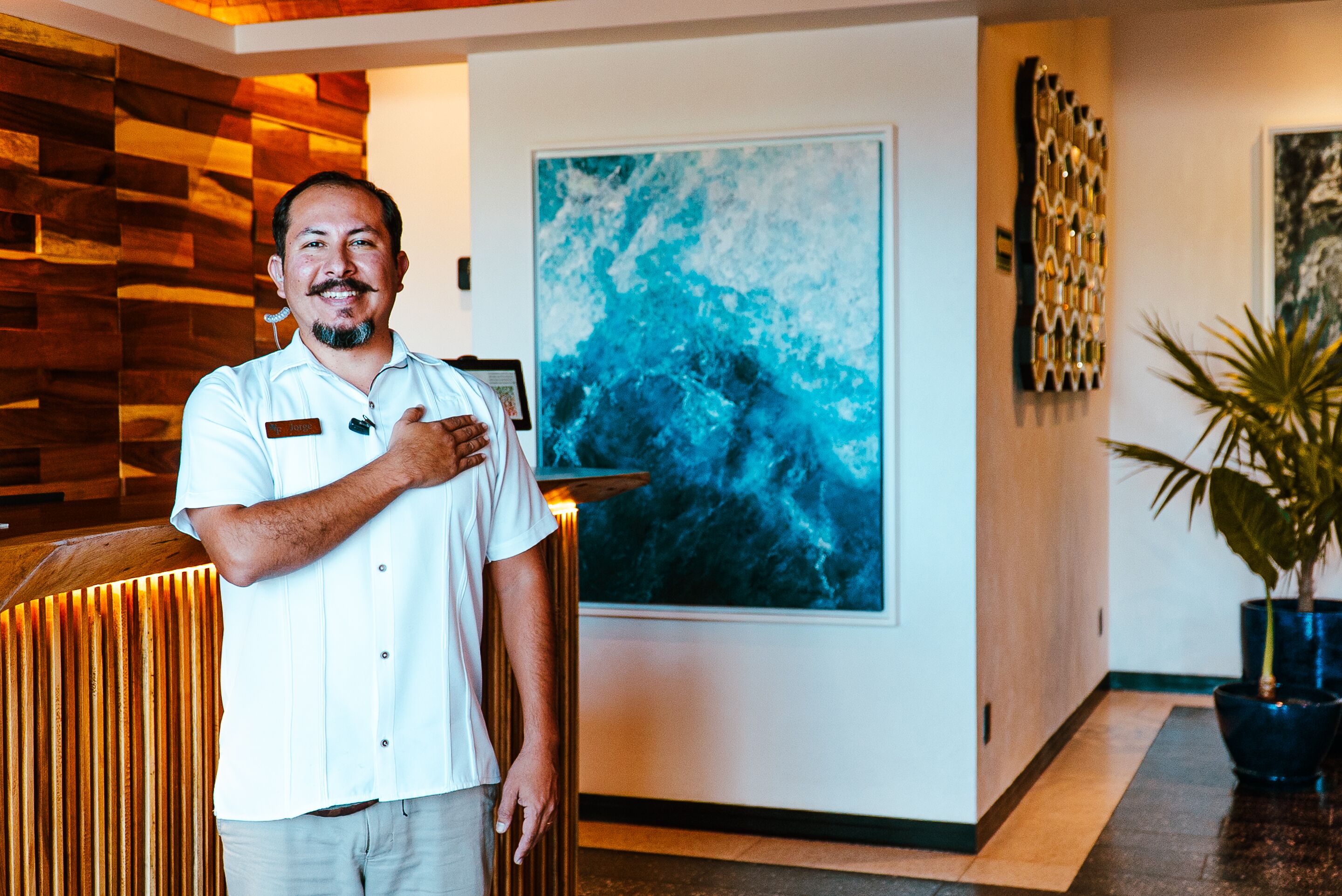 Un homme se tient dans un hall d’entrée, devant un comptoir de réception en bois, un grand tableau abstrait et un pot de fleurs.