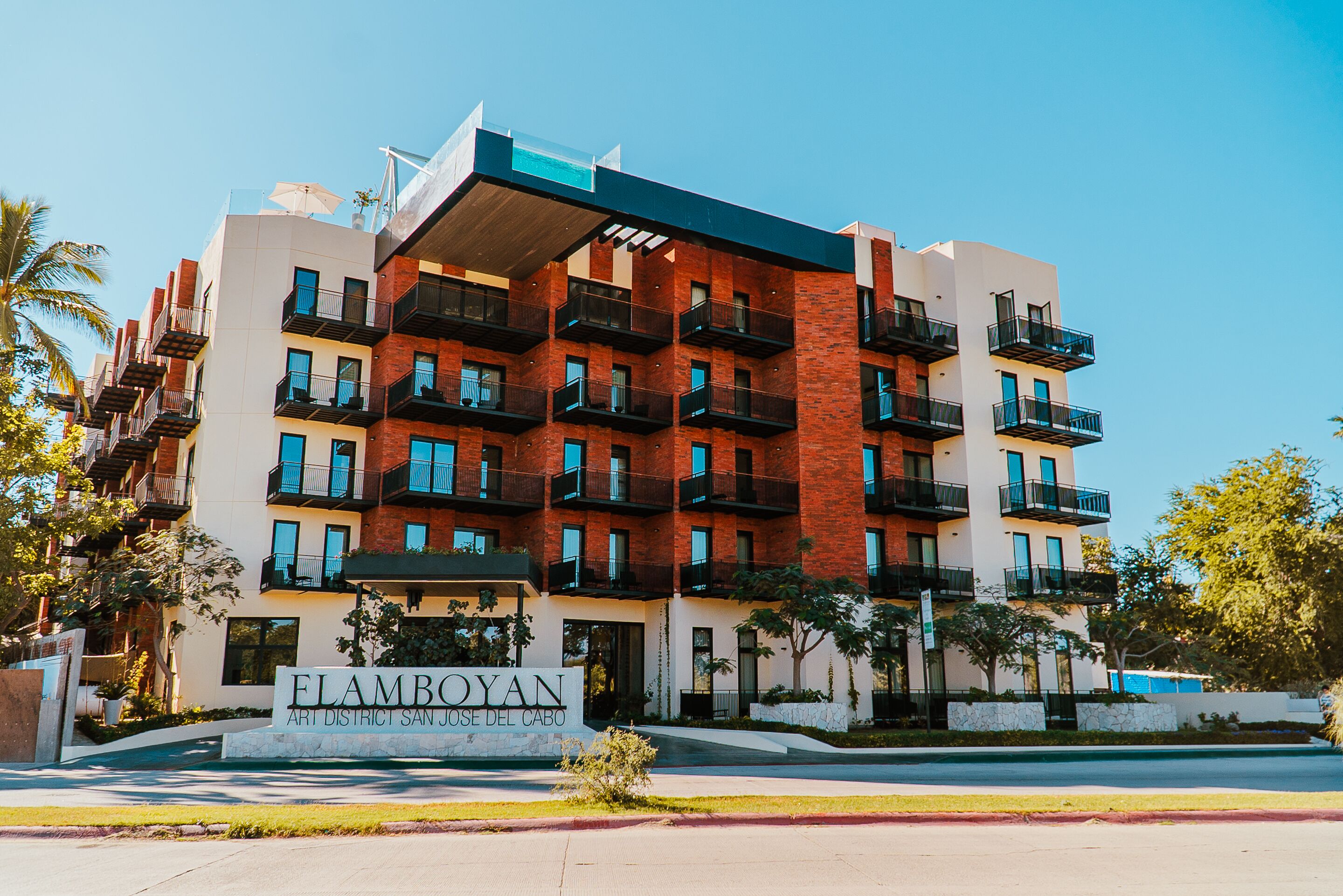 Un immeuble à plusieurs étages doté de balcons, une enseigne indiquant « FLAMBOYAN ART DISTRICT SAN JOSE DEL CABO », et un ciel d’un bleu limpide.