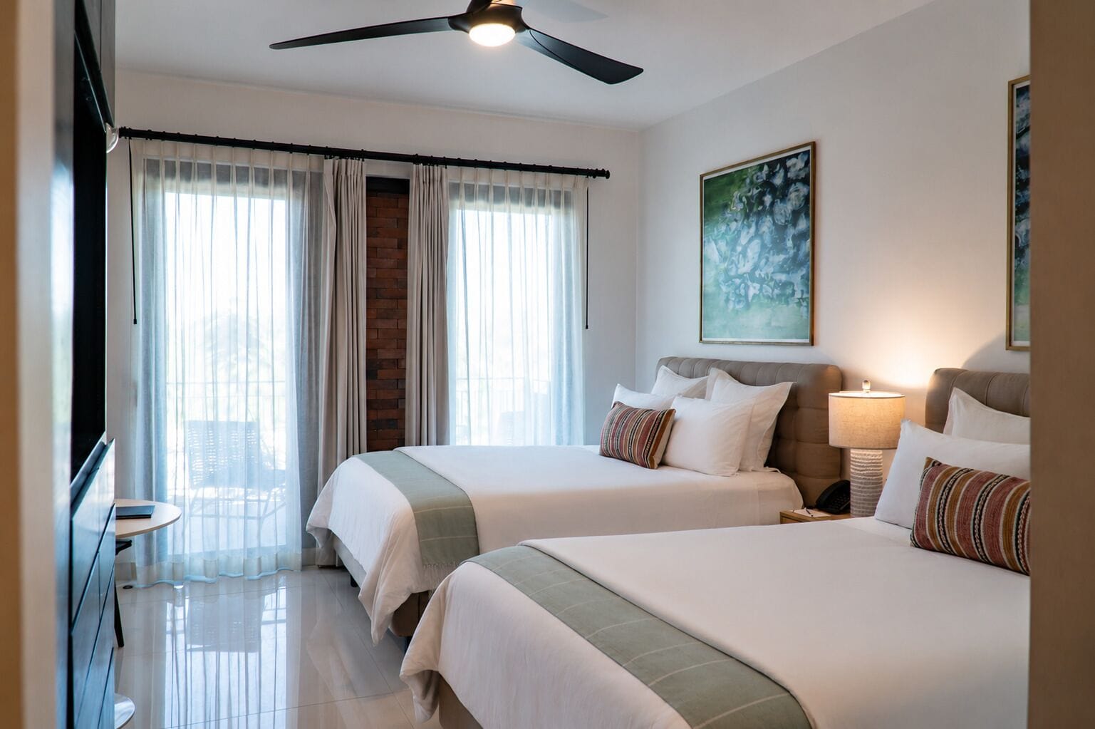 A hotel room with two beds, a ceiling fan, a painting on the wall, and a window with curtains.