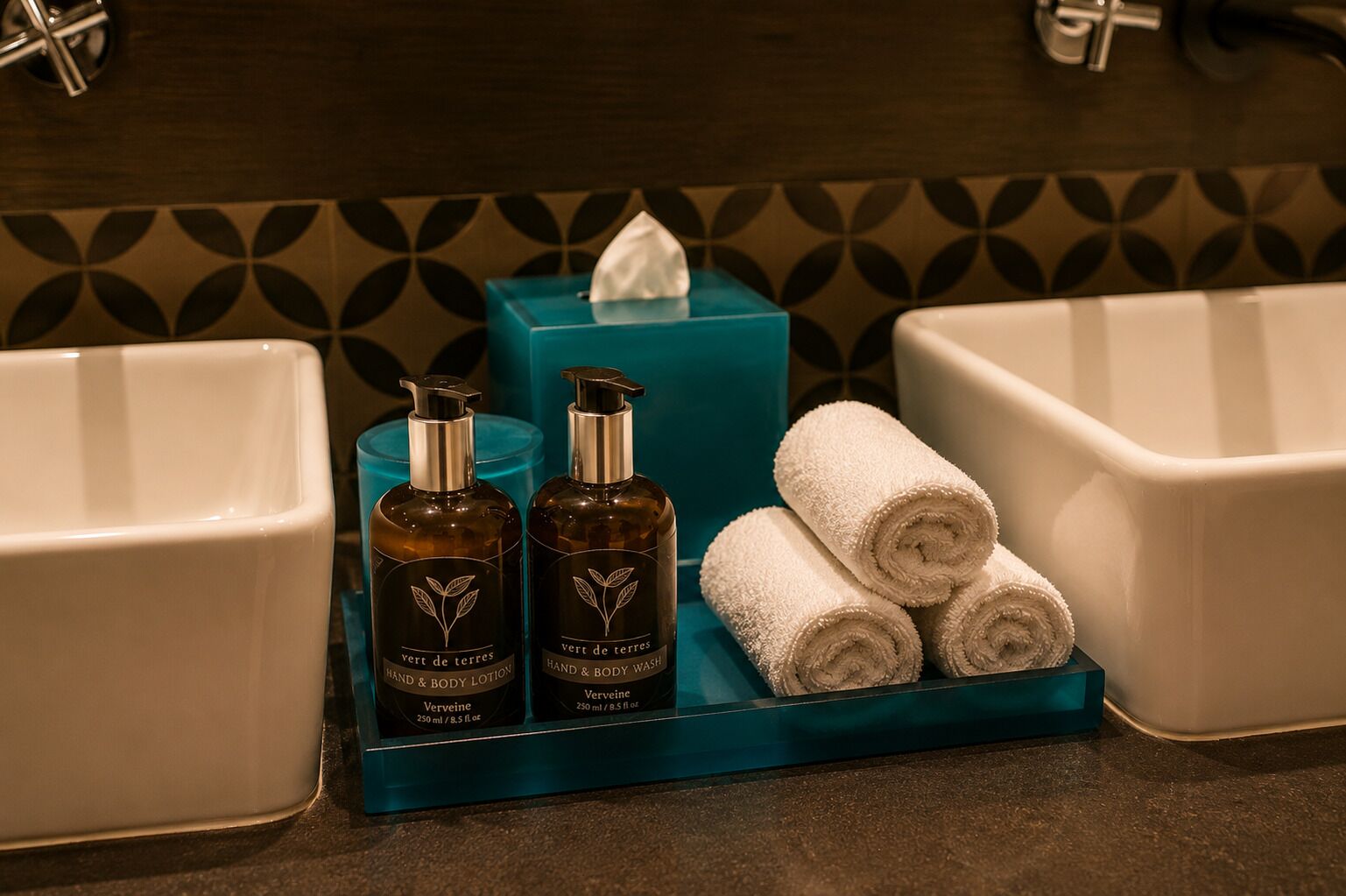 A bathroom vanity with a blue soap dish, two bottles of hand and body wash, and three rolled towels.