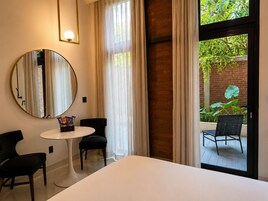 Hotelkamer met een bed, twee stoelen, een tafeltje, een ronde spiegel en uitzicht op groen door een glazen deur.