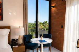 Habitación de hotel con cama, mesita de noche con lámpara, mesa con dos sillas y vista a palmeras desde un balcón.