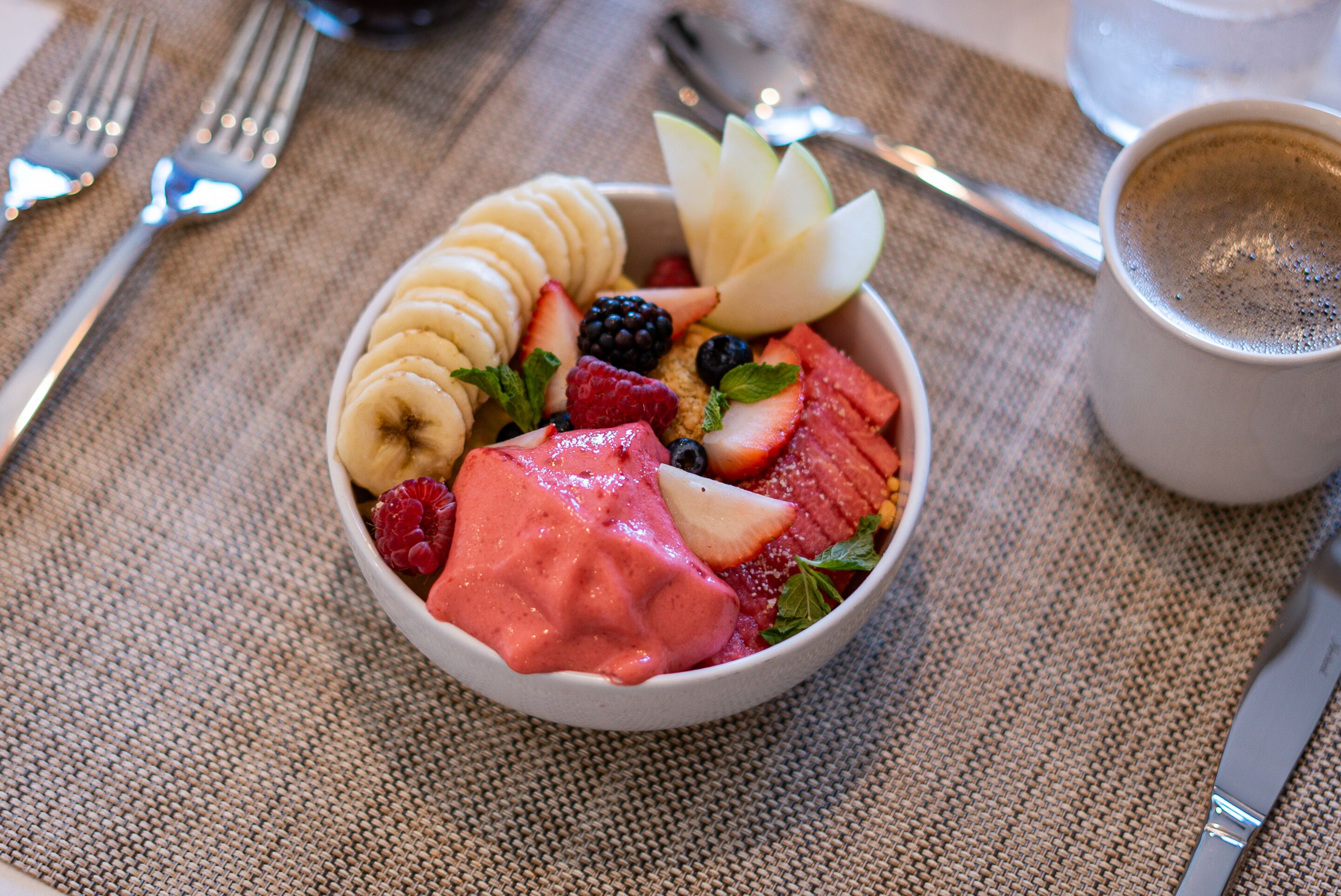 A bowl of fruit salad with strawberries, bananas, and blueberries, accompanied by a cup of coffee on a table with a placemat.