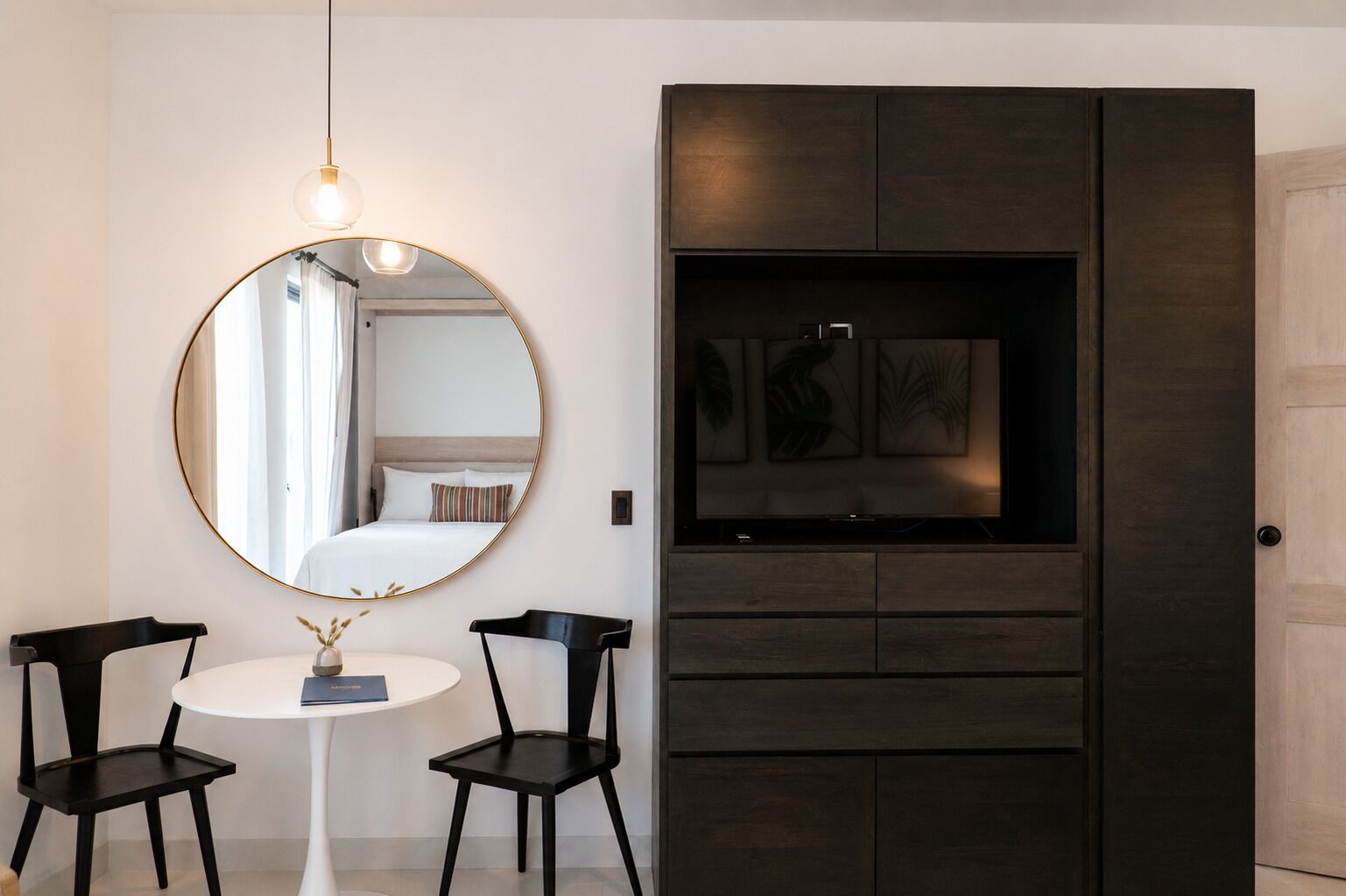 Una habitación con un espejo redondo, una cama con ropa de cama blanca, una mesita con dos sillas y un mueble de madera oscura con obras de arte.