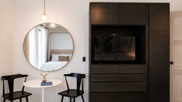 Una habitación con un espejo redondo, una cama con ropa de cama blanca, una mesita con dos sillas y un mueble de madera oscura con obras de arte.