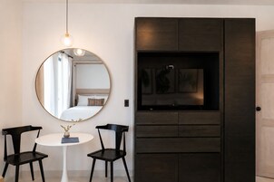 Een kamer met een ronde spiegel, een bed met witte lakens, een kleine tafel met twee stoelen, en een donkere houten kast met kunstwerk.