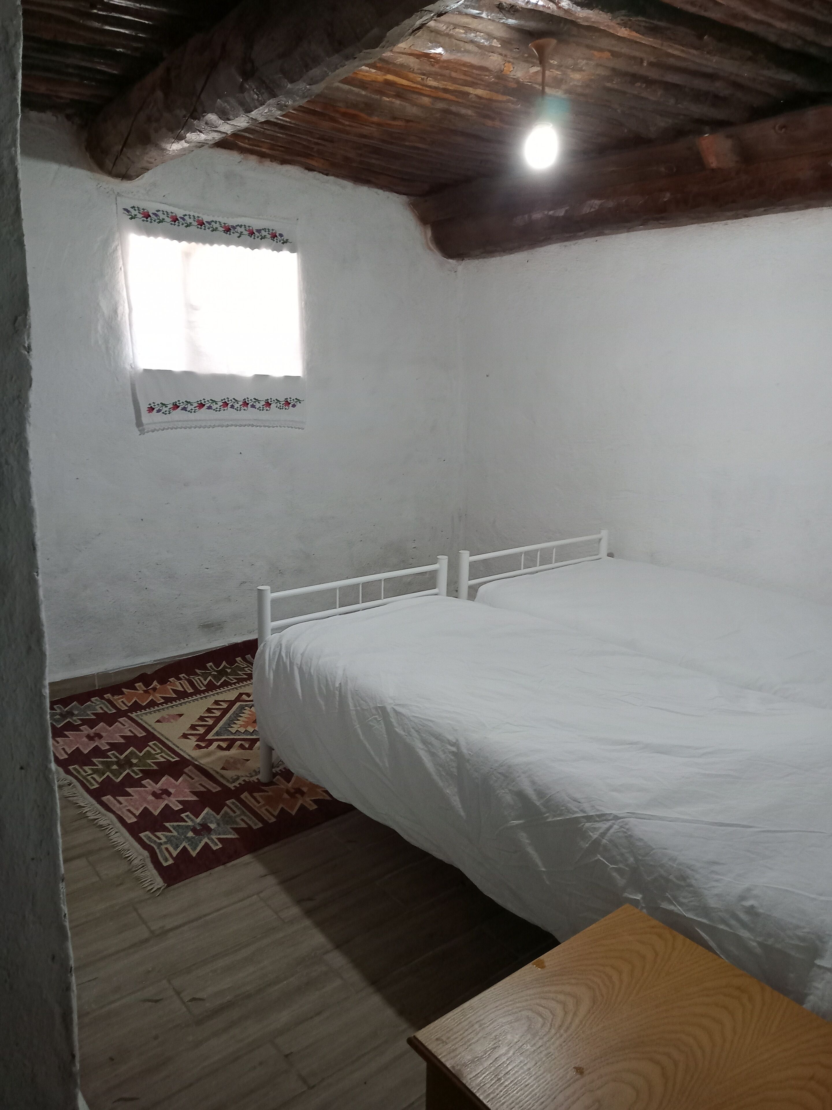 A bedroom with a wooden ceiling, a single bed, a small window with a floral curtain, and a patterned rug.