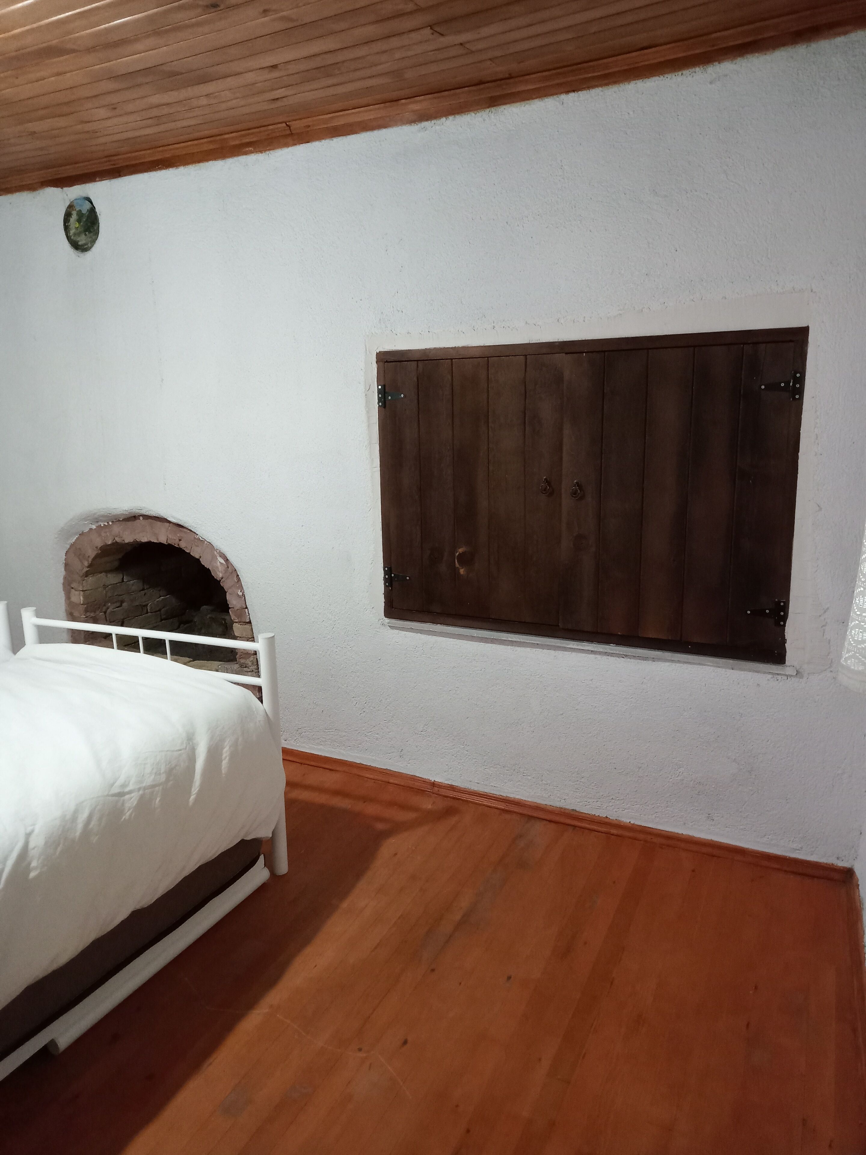 Un dormitorio con suelo de madera, paredes blancas, una cama con ropa de cama blanca y una pequeña chimenea de ladrillo.