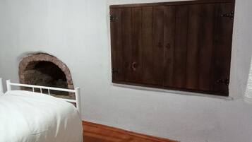 Un dormitorio con suelo de madera, paredes blancas, una cama con ropa de cama blanca y una pequeña chimenea de ladrillo.