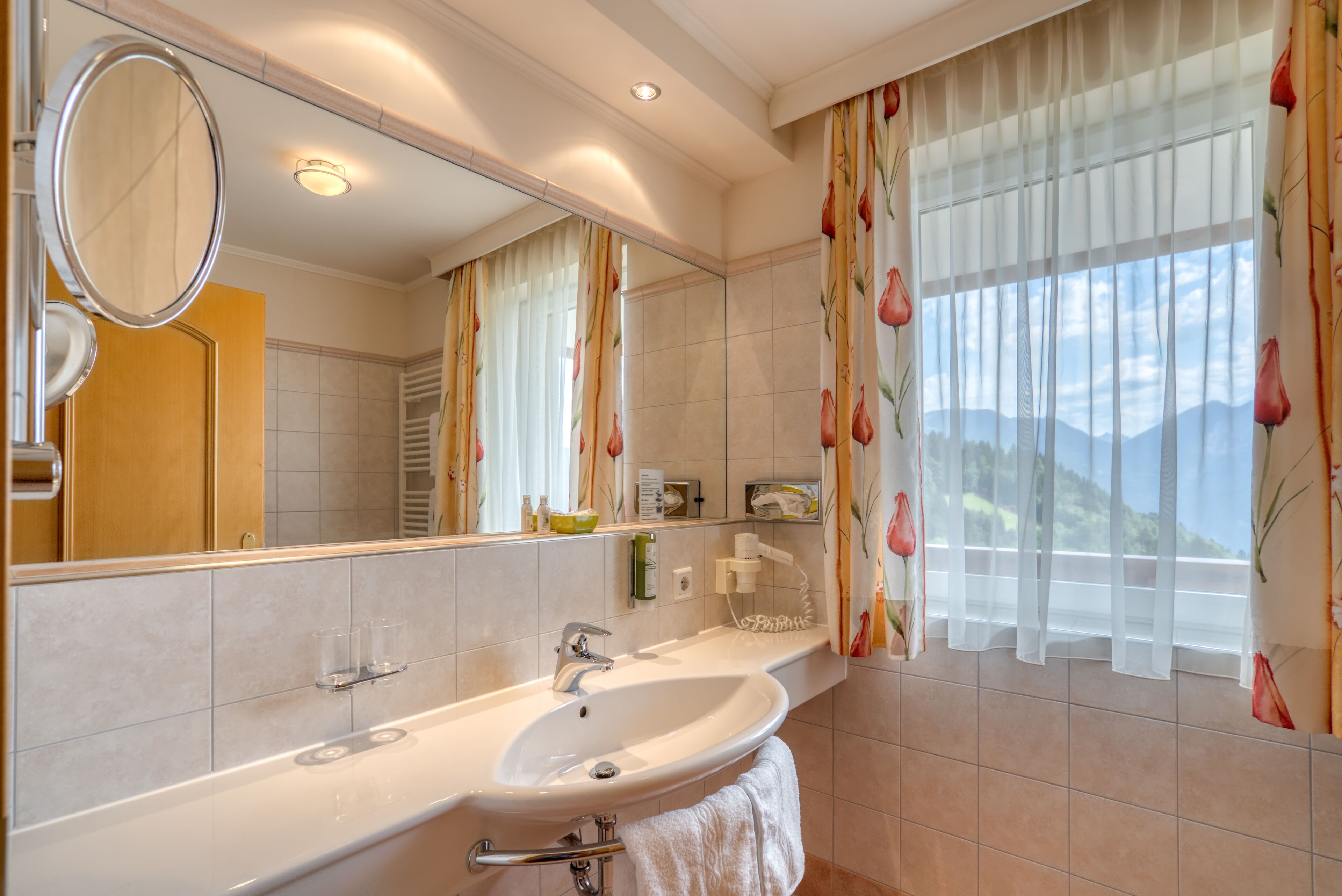 A bathroom with a large mirror, a sink, a towel rack, and a window with floral curtains.