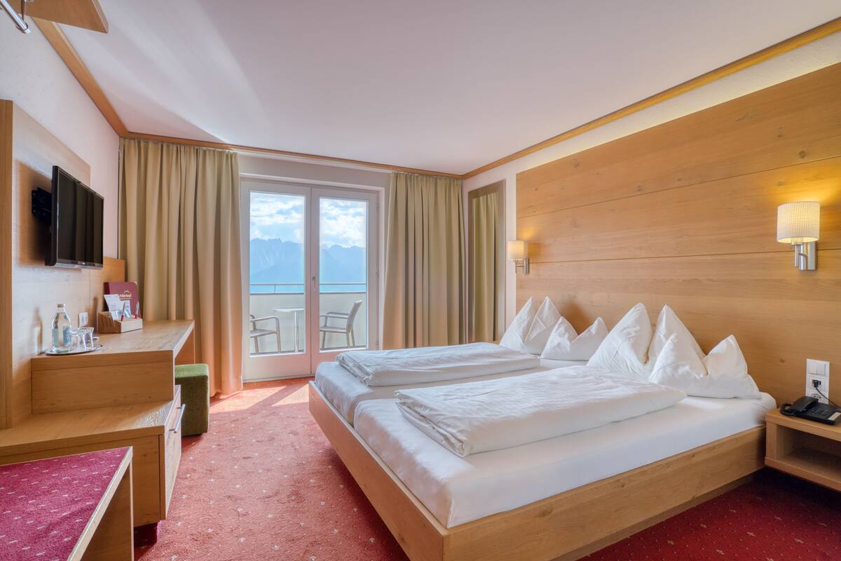 A hotel room with two beds, a desk, a TV, and a balcony with a view of the mountains.