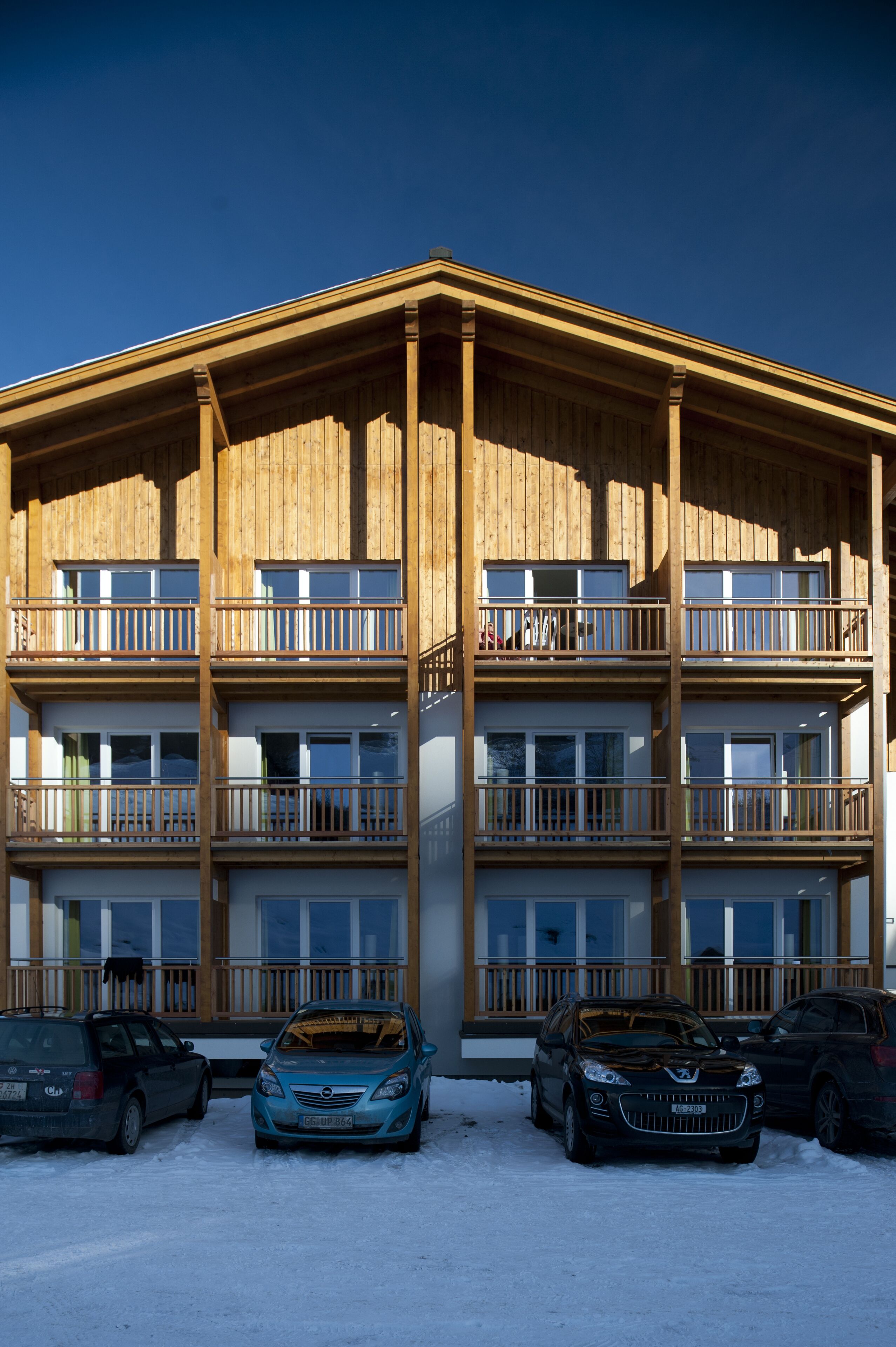 A multi-story building with balconies and a parking area with cars.