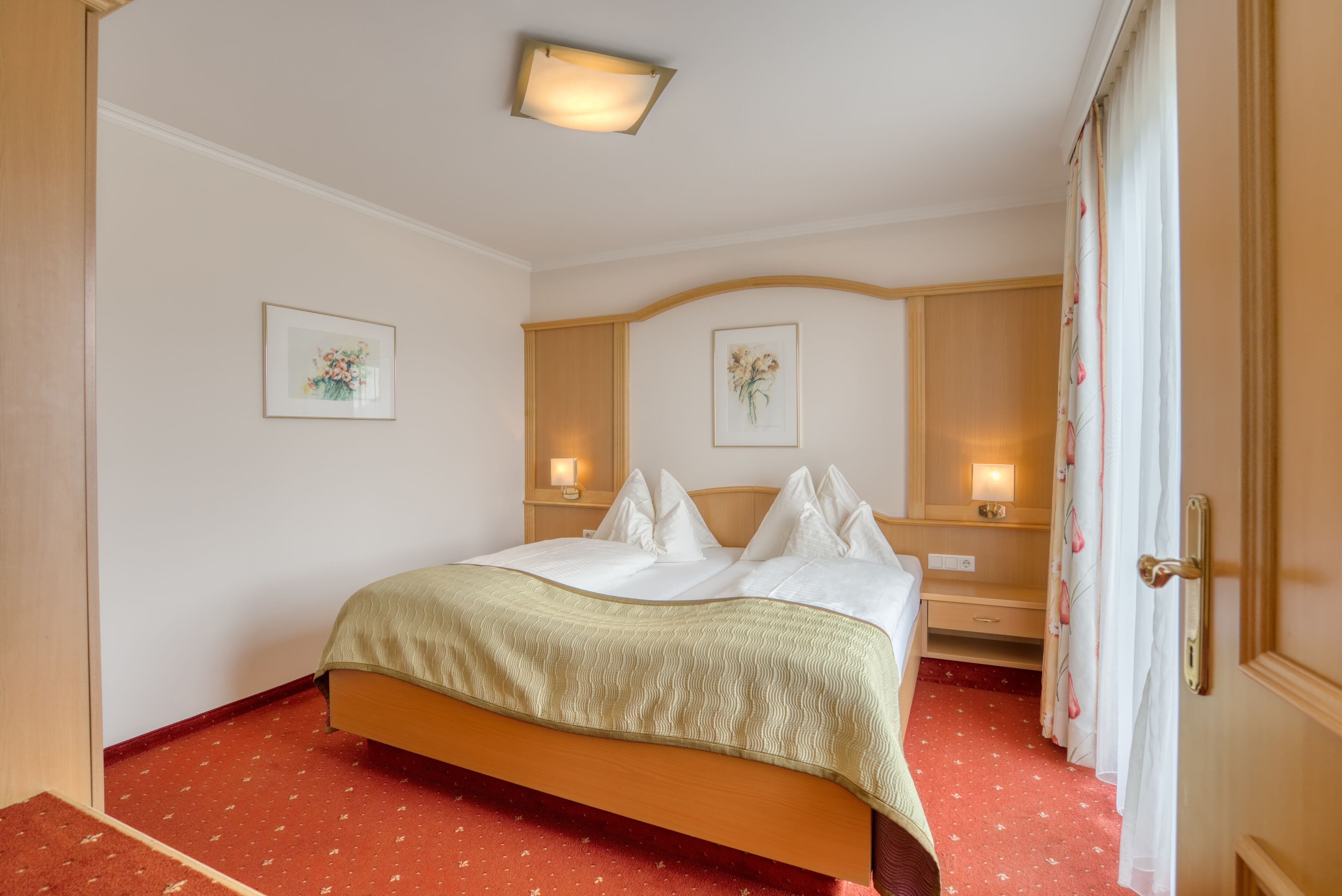 Una habitación de hotel con una cama, dos mesitas de noche, un velador, una lámpara de pared y una puerta con cortina.