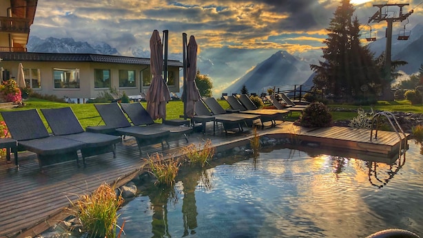Un área de piscina con sillones, una terraza de madera y montañas al fondo.