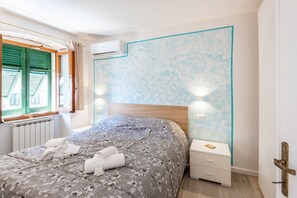 Ein Schlafzimmer mit Bett, Nachttisch, Wandklimaanlage, Fenster mit Rollläden und Heizkörper.