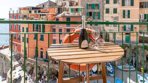 A wooden table with a bottle and glasses on a balcony overlooking a coastal town with colorful buildings and boats. - GuestHost - BOOK NOW! Cà da Mirela x4 WONDERFUL 5TERRE (Riomaggiore)