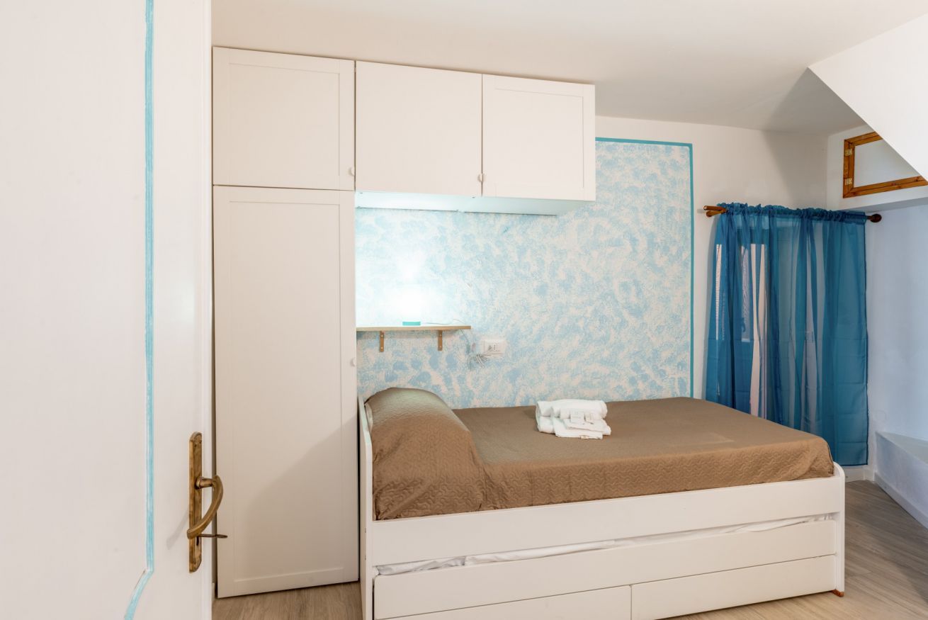 A bedroom with a bed, a wardrobe, a small shelf, and a window with curtains.