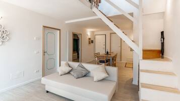 A modern living room with a white sofa, wooden staircase, and a dining area.
