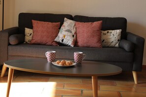 A dark grey sofa with patterned cushions and a round wooden coffee table with two mugs and a plate of cookies.