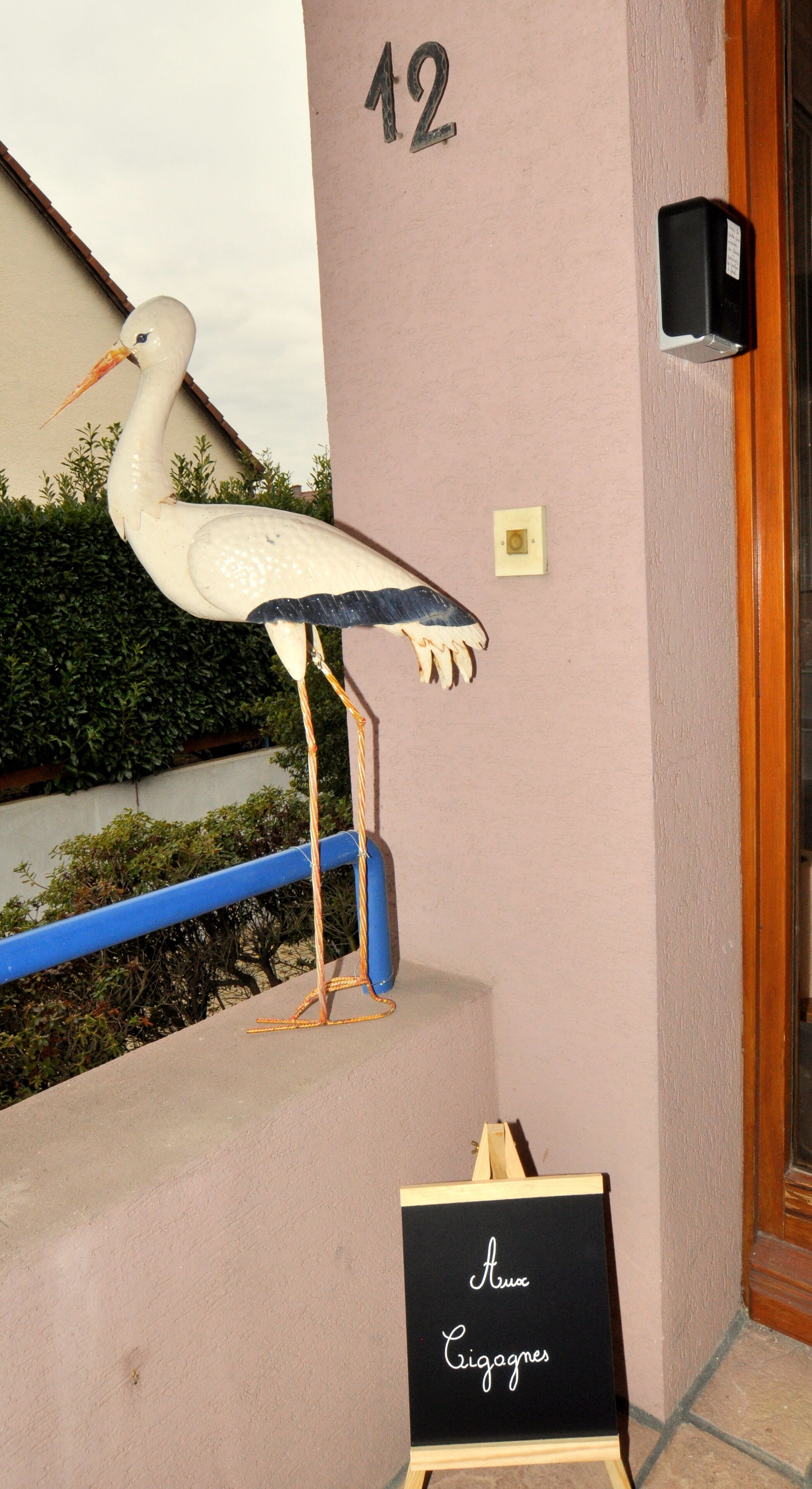 Um edifício com o número '12' na parede, um corrimão azul, uma escultura de pássaro branco e uma lousa com 'Amo' e 'Cigagnes' escritos.