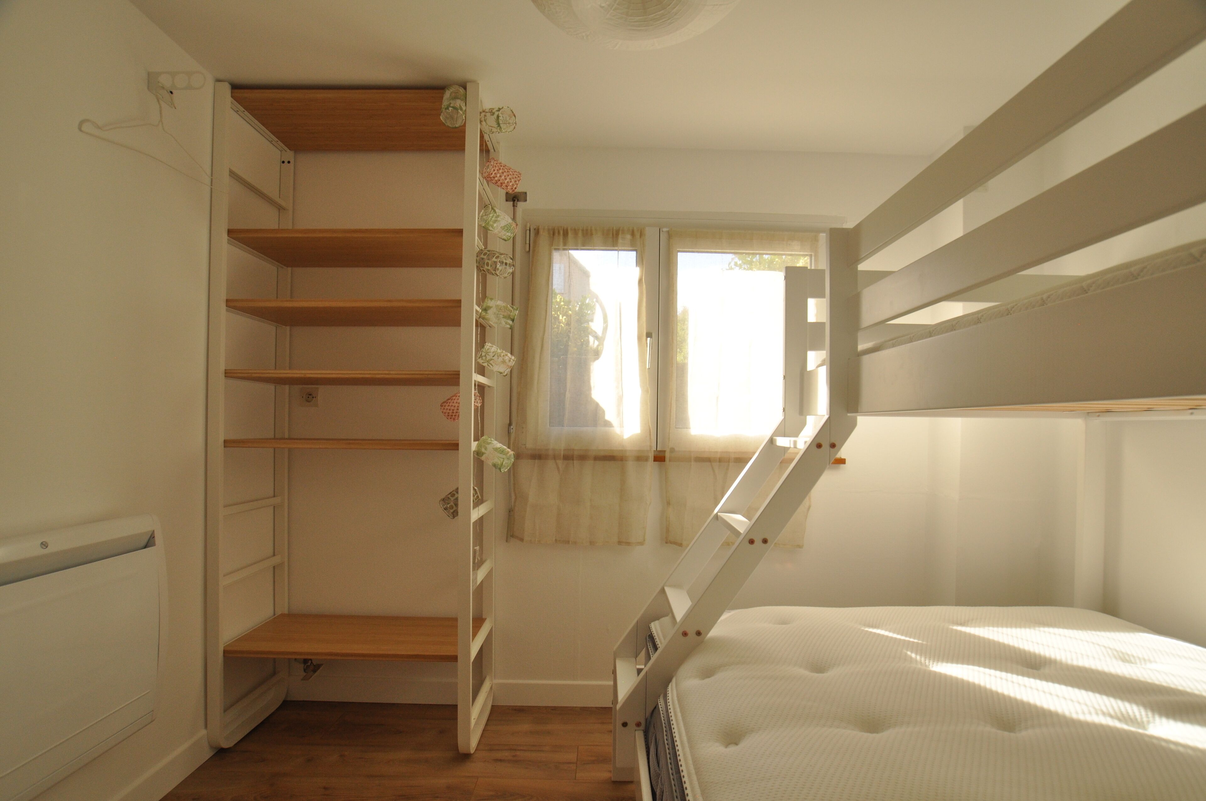 Une chambre à coucher de taille réduite, avec un lit superposé, une petite fenêtre avec des rideaux et une étagère en bois.