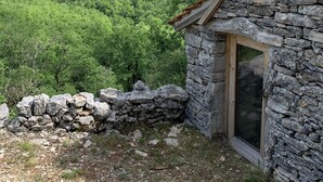 A stone cottage with a wooden door, surrounded by a stone wall and a stone roof, set against a backdrop of lush green hills and a clear blue sky. - L'Hermitage, an old stone shepherd's hut (Salvagnac-Cajarc)