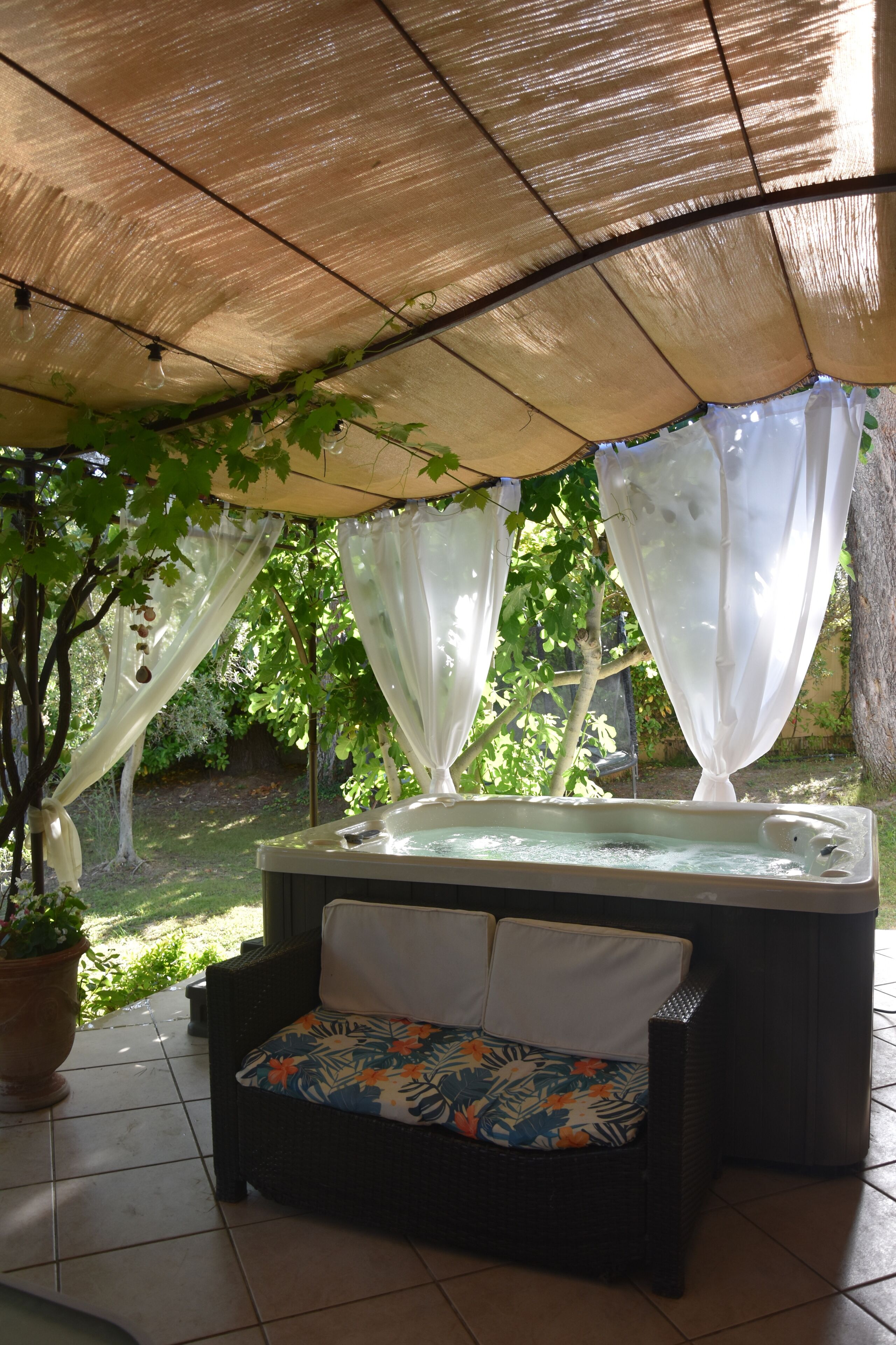 A hot tub under a shaded area with a wicker sofa and a potted plant.