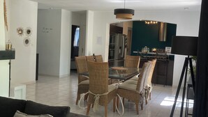 Un salon moderne avec un canapé aux couleurs sombres, une table à manger en verre entourée de chaises en osier, et un coin cuisine à l’arrière-plan.